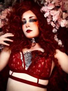 Sensual glamour portrait photography featuring long, curly red hair, red lace lingerie, and a backdrop of beautiful flowers.